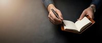 Professional businessperson writing in open notebook with pen at dark desk, planning and note-taking for productivity and organization in modern workspace.