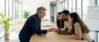 Mature male executive in dark suit joining hands with diverse female team members at wooden table in bright office space, symbolizing unity, teamwork, and corporate collaboration.