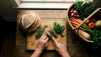 Fresh herbs being chopped on rustic wooden cutting board with garden vegetables in wicker basket. Organic cooking preparation scene for healthy recipes.