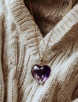 Purple crystal heart pendant on gold chain against cozy knit sweater background, romantic jewelry detail in warm neutral tones with soft lighting.