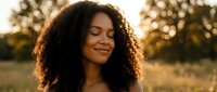 Young African American woman with curly hair smiling peacefully with eyes closed in warm golden hour sunlight outdoors for wellness and self-care concepts.