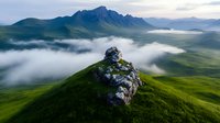 Dramatic mountain landscape with morning fog rolling through green valleys, rocky outcrop on grassy hill foreground against majestic peaks in distance. Atmospheric nature scene.