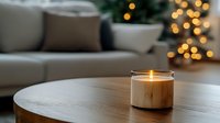 Glowing candle in wooden holder on coffee table creates cozy atmosphere with blurred Christmas lights and modern sofa in background, warm home interior setting.