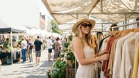 Woman in sunglasses and straw hat browsing clothing at outdoor market or street fair, shopping for summer fashion and casual wear.