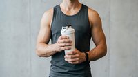 Muscular man with tan skin wearing dark sleeveless shirt holding protein shake bottle for fitness, nutrition, and workout content.