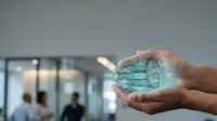 Young adult male hand holding glowing turquoise digital city model in modern office environment with blurred colleagues in background, representing urban development and technology innovation.