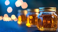 Glass jar with golden honey or syrup on wooden surface against blurred blue background with glowing bokeh lights creating magical evening atmosphere.
