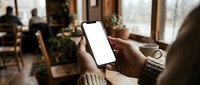 Person holding smartphone with blank white screen in cozy cafe setting. Mockup template for mobile app presentation and digital marketing materials.