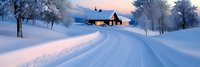 Snowy winter road leading to cozy cabin with warm lights at twilight, surrounded by frost-covered trees in peaceful countryside landscape.