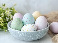 Pastel colored Easter eggs with polka dot patterns and speckled designs arranged in decorative ceramic bowl against light background with spring greenery.