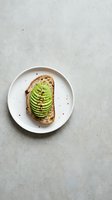 Sliced avocado on toasted bread served on white plate with red pepper flakes, shot from above on concrete surface for food styling and culinary content.