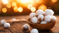 Raw cotton balls in wooden bowl on rustic table with warm bokeh background, natural organic textile material in soft evening light.