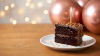 Chocolate birthday cake slice with lit candle on plate, rose gold balloons and bokeh lights creating festive celebration atmosphere for party occasions.