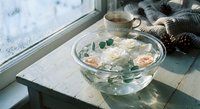 Elegant glass bowl with floating roses and eucalyptus leaves on wooden windowsill, cozy home spa relaxation setup with warm blanket and tea cup.