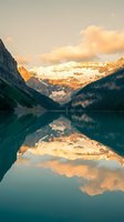 Serene mountain lake with snow-capped peaks reflecting in calm turquoise water during golden hour sunrise creating mirror symmetry in pristine wilderness landscape.
