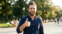 Happy Caucasian businessman with beard giving thumbs up gesture in sunny park setting during golden hour for positive business and success concepts.