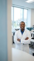 Confident African American male scientist in white lab coat standing with crossed arms in modern research laboratory with city view windows.