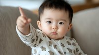 Asian baby boy in bear print pajamas pointing upward while sitting on gray couch, expressing curiosity and wonder with innocent facial expression.