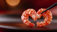 Cooked pink shrimp held with black chopsticks forming heart shape against dark red background with soft bokeh lighting, macro food photography.