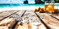 Melting ice cubes on wooden deck near tropical beach with cocktail and orange slices, turquoise ocean and blue sky in background, summer vacation concept.