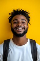 Young African American man with dreadlocks and beard wearing white t-shirt and backpack, smiling genuinely against vibrant yellow background. For lifestyle and diversity campaigns.
