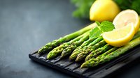 Fresh green asparagus spears with lemon halves and mint leaf on dark stone board against blurred background. Food photography for healthy eating concept.