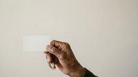Male hand with dark skin tone holding blank white card against neutral beige background for business, marketing, advertising, and promotional messaging purposes.