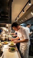 Professional chef plating gourmet dishes in modern commercial kitchen with track lighting and stainless steel equipment for restaurant service.