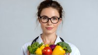 Young female nutritionist in white coat and glasses holding fresh vegetables and fruits in bowl against light background, promoting healthy eating habits.