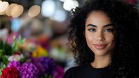 Young African American woman with natural curly hair smiling warmly at camera, surrounded by vibrant colorful flowers in soft bokeh background. Positive authentic expression.