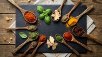 Colorful spices and herbs arranged in wooden spoons on dark slate board. Fresh basil, turmeric, paprika, oregano create culinary cooking ingredient display.