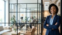 Young African American female professional wearing navy blazer and light blue shirt, standing with arms crossed in modern office corridor with glass partitions and colleagues visible in background, representing corporate leadership, business confidence, and workplace diversity.