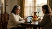 Senior Caucasian woman with glasses and young adult woman collaborating on tablet computer at wooden dining table in cozy home interior setting.