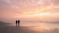 Romantic couple silhouettes walking hand in hand on peaceful beach during golden sunset with soft pink and orange sky reflecting on wet sand creating serene moment.