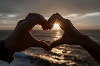 Hands forming heart shape silhouette against golden sunset over ocean water. Romantic love symbol gesture at beach during golden hour for Valentine's Day concepts.