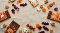 Halloween chocolate bars with pumpkin faces and assorted candies arranged in circle on beige background. Festive autumn sweets for trick or treat celebration.