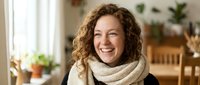 Happy young Caucasian woman with curly hair wearing cozy knit scarf smiling warmly in bright modern home interior with plants and natural light.