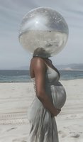 Pregnant African American woman in flowing white dress stands on beach with transparent sphere creating surreal portrait effect against ocean backdrop.