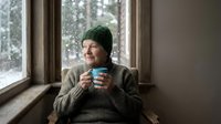 Senior Caucasian man in green knit hat enjoying hot beverage by window during winter, cozy indoor moment with warm lighting and peaceful atmosphere.