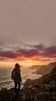 Hiker with backpack standing on rocky cliff overlooking ocean at dramatic sunset with golden and orange sky, coastal landscape with distant cliffs and waves below.