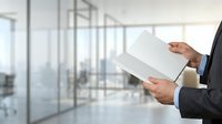 Businessman in dark suit holding blank white tablet or clipboard in modern office with glass partitions and natural light.