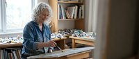 Senior Caucasian woman with gray hair working on laptop at wooden desk in bright home office with bookshelves, representing remote work and mature professional lifestyle.