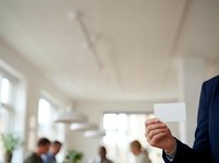 Businessman in dark suit holding blank white card in modern office environment with colleagues and natural light from windows.