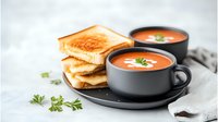 Creamy tomato soup garnished with fresh herbs served in dark ceramic bowls alongside golden grilled cheese sandwiches on light marble background.