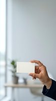 Businessperson holding blank white card in modern office environment with natural light and blurred background, suitable for corporate branding and business card mockups.