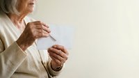Senior woman with gray hair and fair complexion holding blank white card in both hands, wearing beige cardigan, isolated against neutral background for messaging.