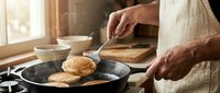 Caucasian man in apron flipping golden pancakes in cast iron skillet on stovetop, warm morning kitchen light, homemade breakfast cooking scene.