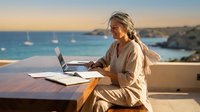 Mature woman with gray hair working on laptop at outdoor table overlooking turquoise sea and sailboats during golden hour sunset, remote work and travel lifestyle concept.