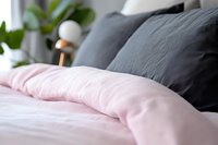 Cozy bedroom interior with soft pink and dark gray bedding, close-up view of rumpled linen pillows and duvet cover against blurred background with houseplant.