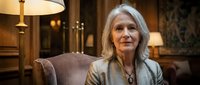 Elegant senior Caucasian woman with gray hair sitting in a luxurious classic interior, exuding confidence and sophistication in warm ambient light.